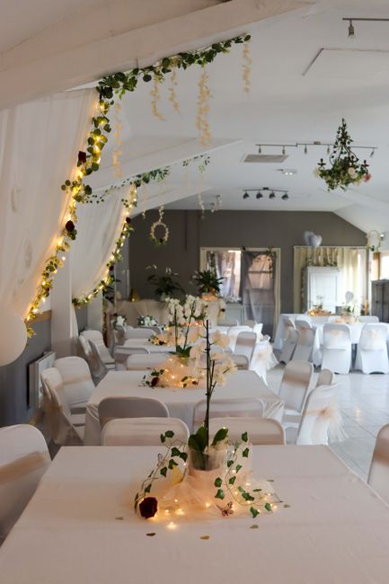 The image depicts an elegantly decorated event space with numerous round banquet tables covered in white tablecloths, each adorned with floral arrangements including white roses and greenery in clear