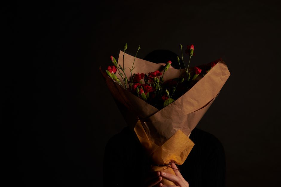 A person holding a large bouquet of fresh red and pink flowers, including roses and lisianthus, wrapped in brown kraft paper and tied with a dark ribbon. The bouquet is positioned in front of their fa