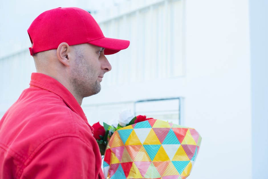 A delivery person wearing a red cap and red jacket holds a vibrant, multicolored floral gift wrapped in patterned paper with geometric shapes, standing outdoors near a modern light-colored building. T