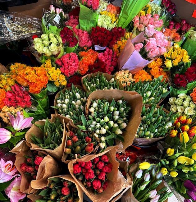 A vibrant display of fresh floral bouquets arranged closely together, featuring a variety of flowers including tulips, roses, lilies, and carnations. The tulips are predominantly white and red, wrappe